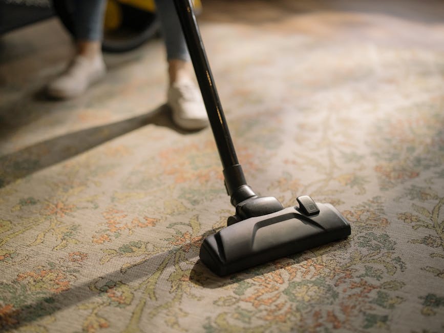 Close-up of a vacuum cleaner nozzle actively cleaning a patterned area rug with intricate floral motifs in muted shades of beige, green, and orange, in a well-lit room. The vacuum attachment is black 