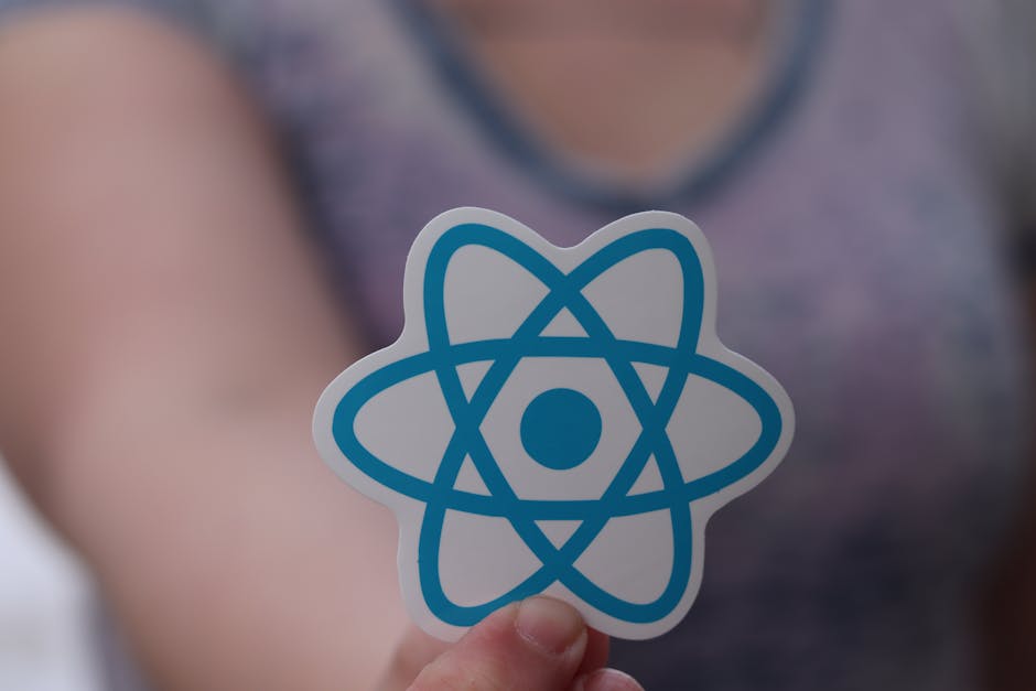 Close-up of a person holding a sticker featuring a blue atom symbol on a white background, with a blurred background that appears to be part of a modern kitchen or office space. The surface nearby inc